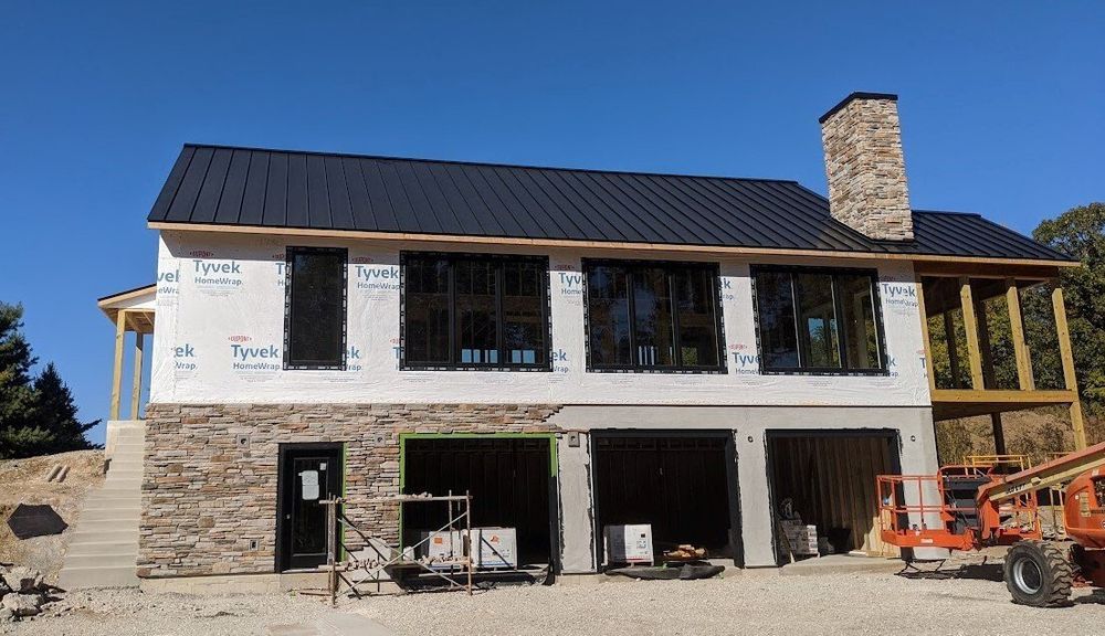 Two-story house under construction with stone facade, black windows, and a black roof against a blue sky.