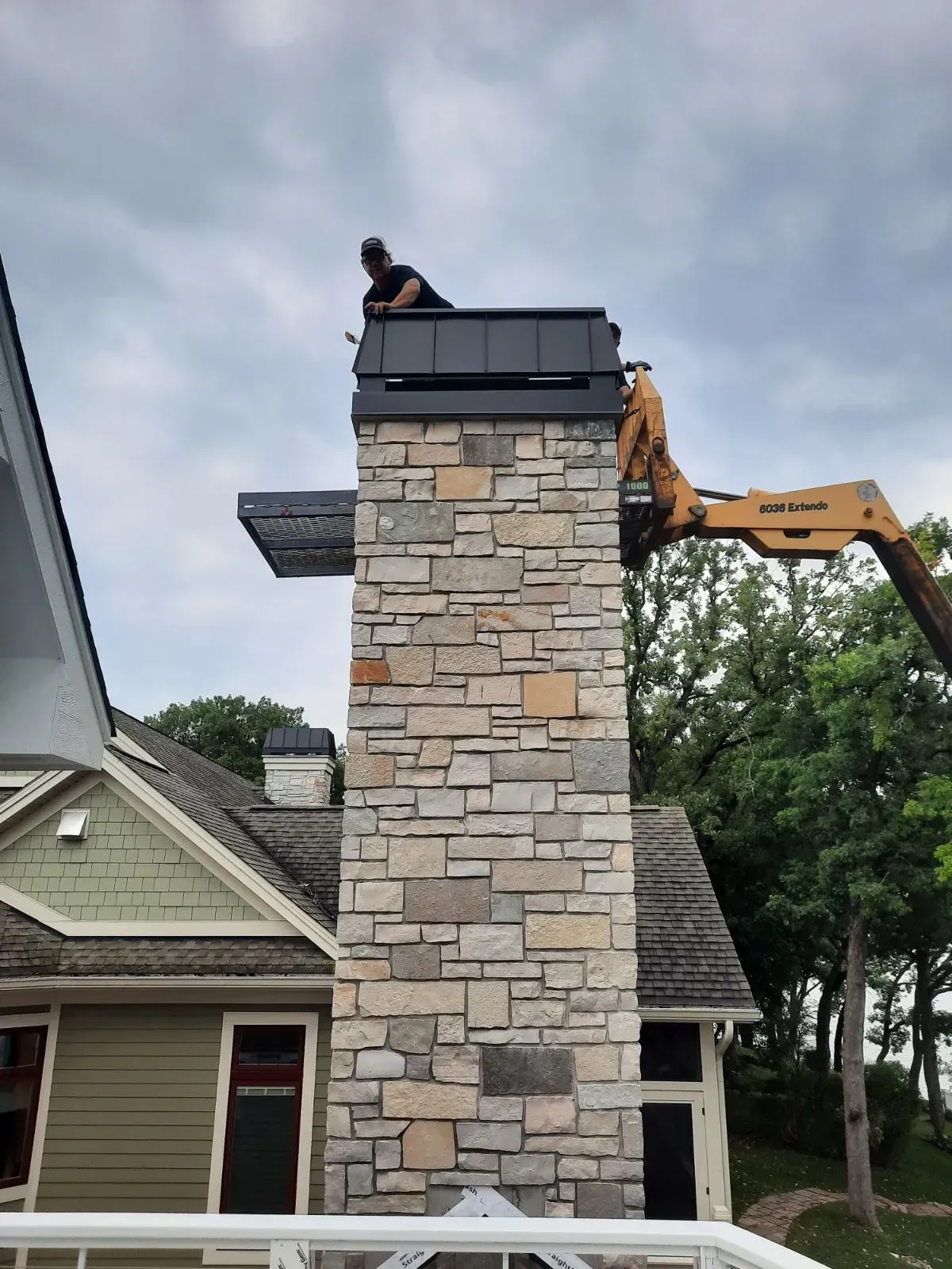 Person on lift working on a stone chimney. Cloudy sky, building in background.