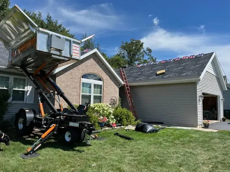 A house with a mechanical lift in the yard, likely for roofing work. Ladder, and debris are visible. Blue sky. A house with a mechanical lift in the yard, likely for roofing work. Ladder, and debris are visible. Blue sky.