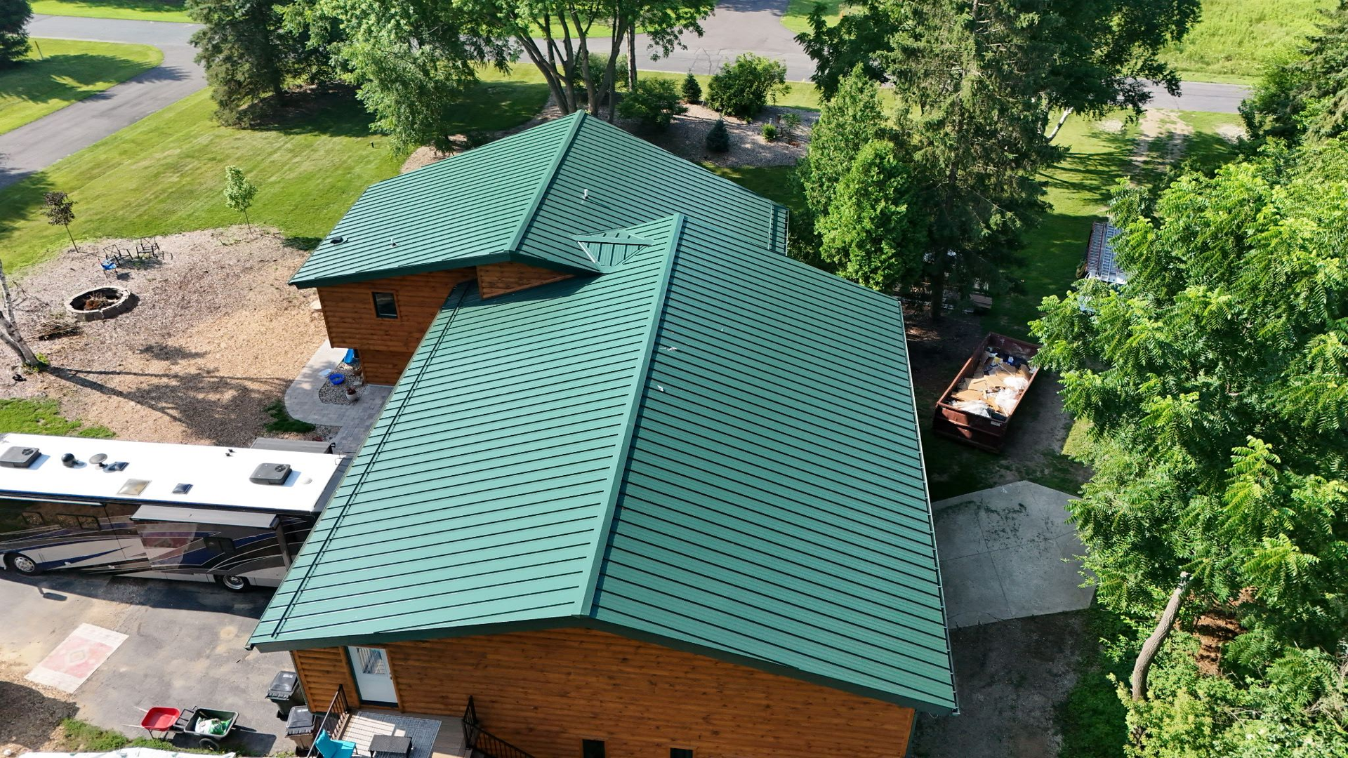 Green metal roof on a wooden building, viewed from above, surrounded by trees and a RV.