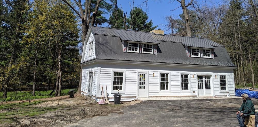 White house with dark roof and dormer windows, surrounded by trees. A person is sitting nearby.