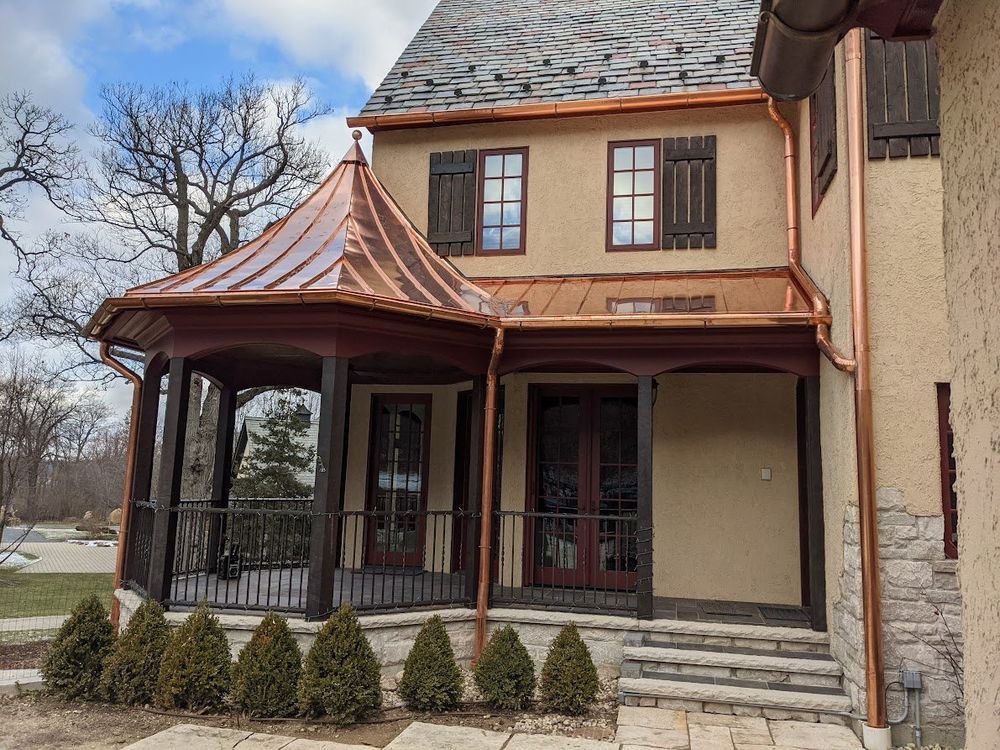 Copper-roofed porch and house with dark shutters. Brown trim, tan walls. Stone steps and a row of green bushes.