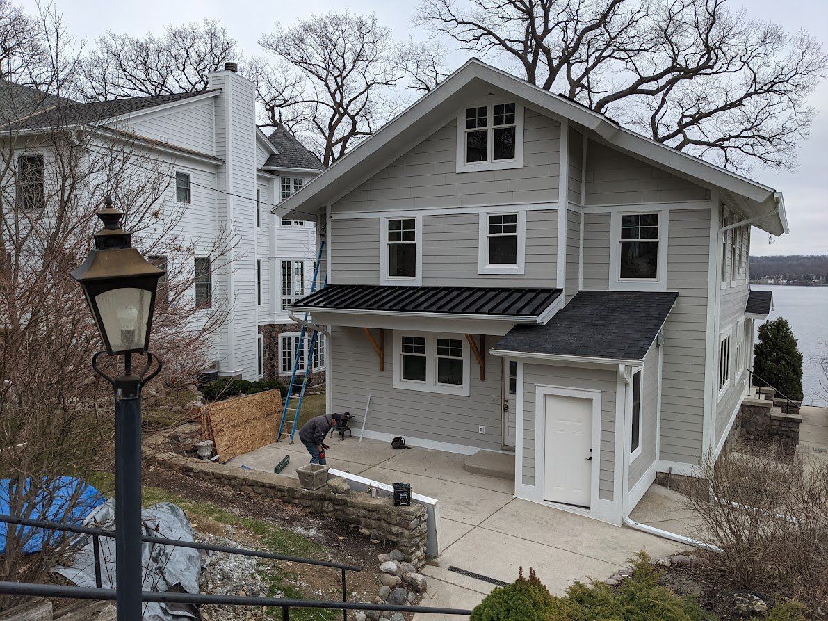 Two-story house with gray siding, white trim, and black roof. A person works in front. Trees and lake are in the background.
