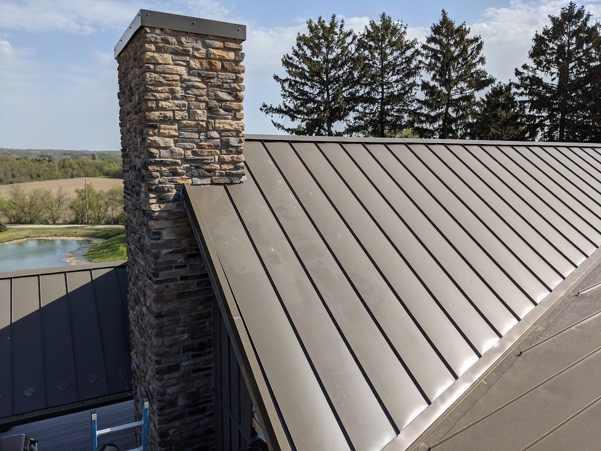 Brown metal roof with a stone chimney against a blue sky with trees and a landscape view.