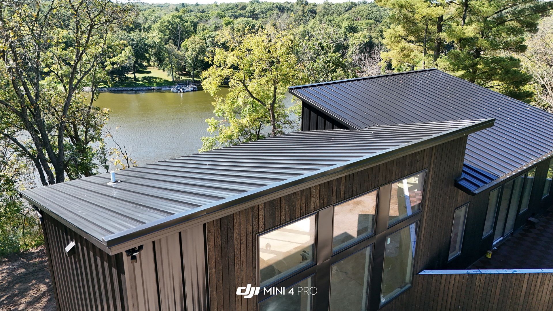 Modern cabin with dark metal roof, large windows, and view of a lake and trees.