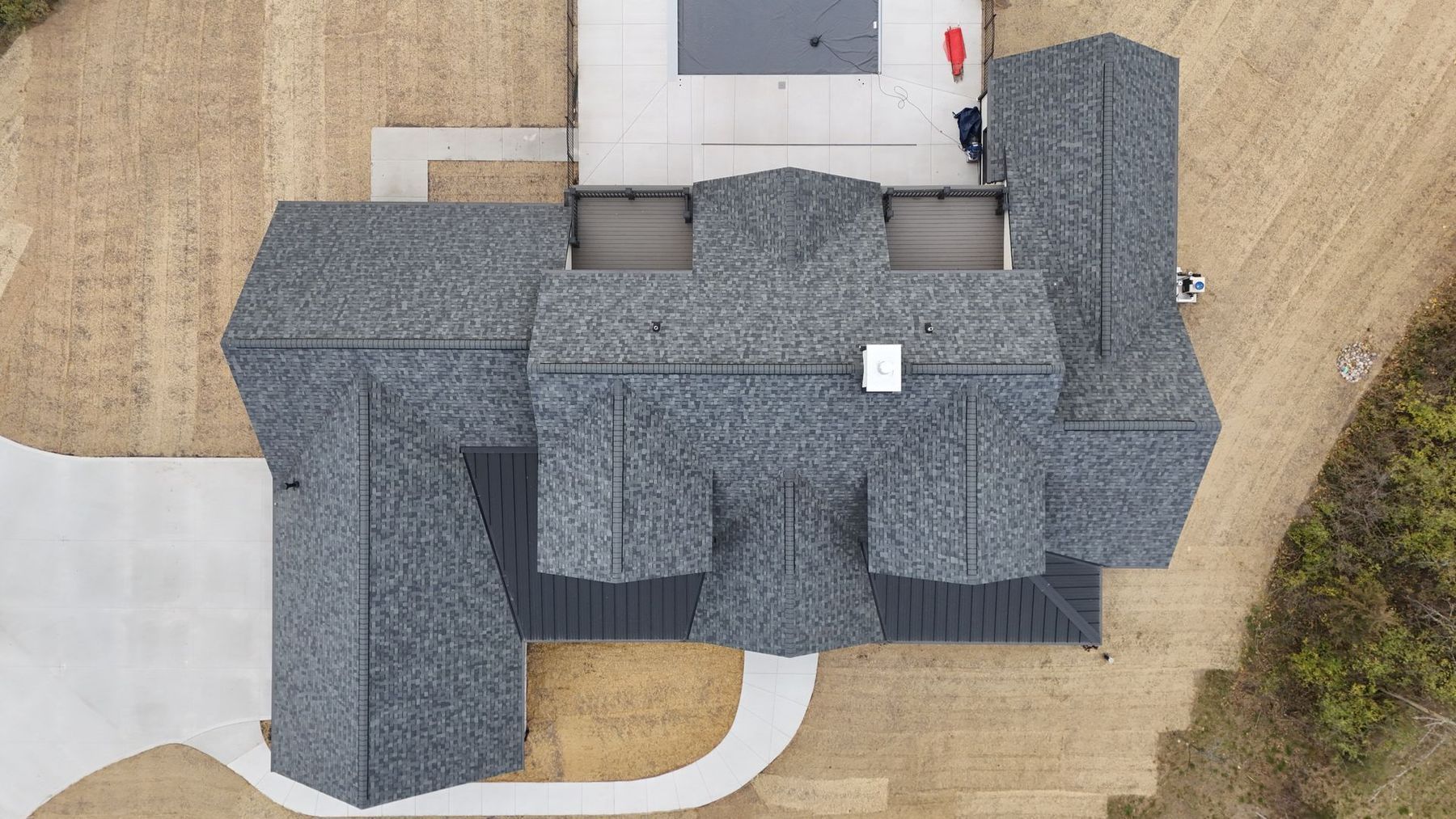 Overhead view of a modern house with a dark gray shingled roof, a pool, and surrounding brown yard.