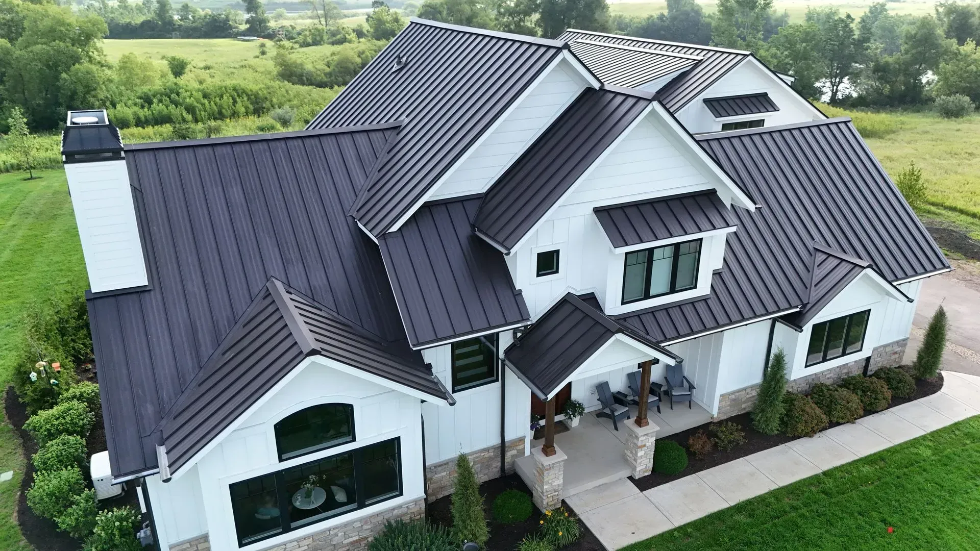 White farmhouse with dark gray metal roof, chimney, and stone accents, set in a green field.