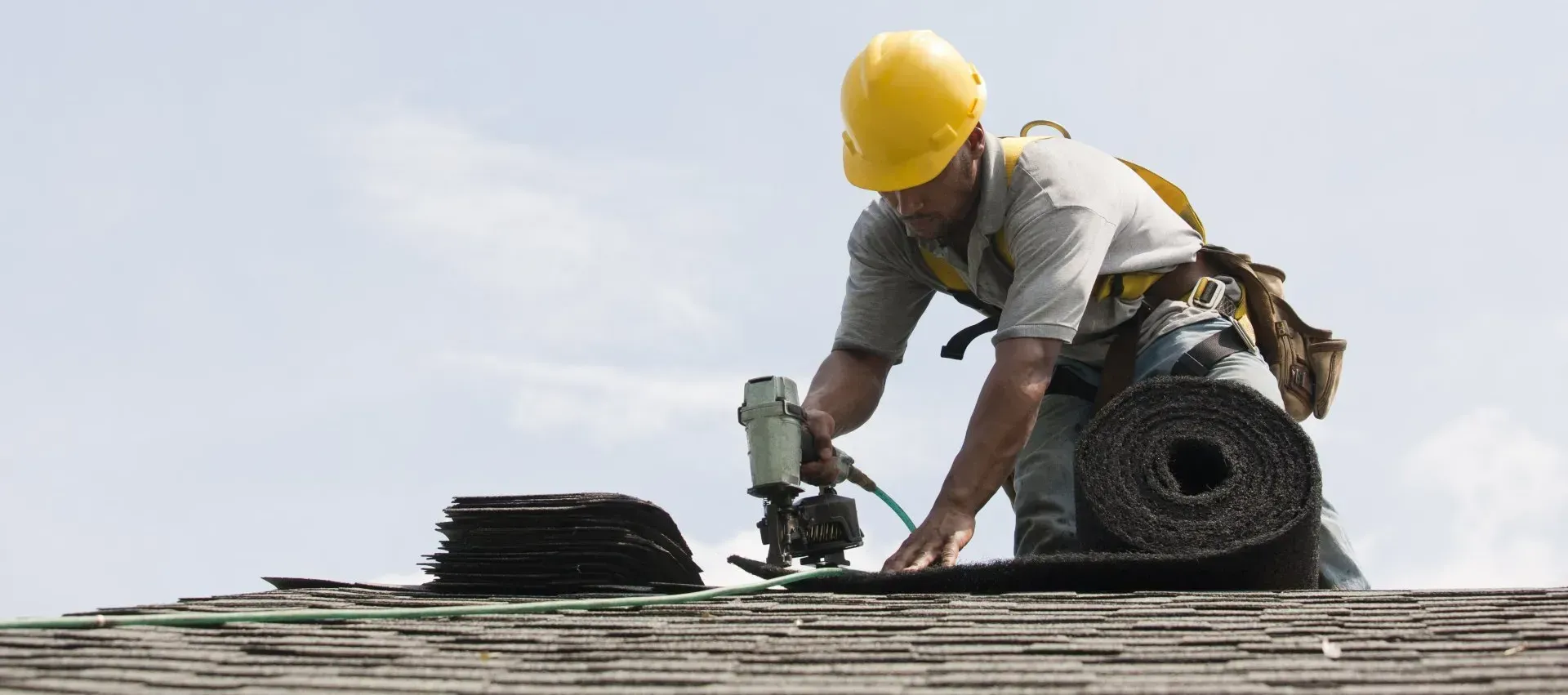 Roofer in yellow hard hat uses a nail gun to install shingles on a rooftop.