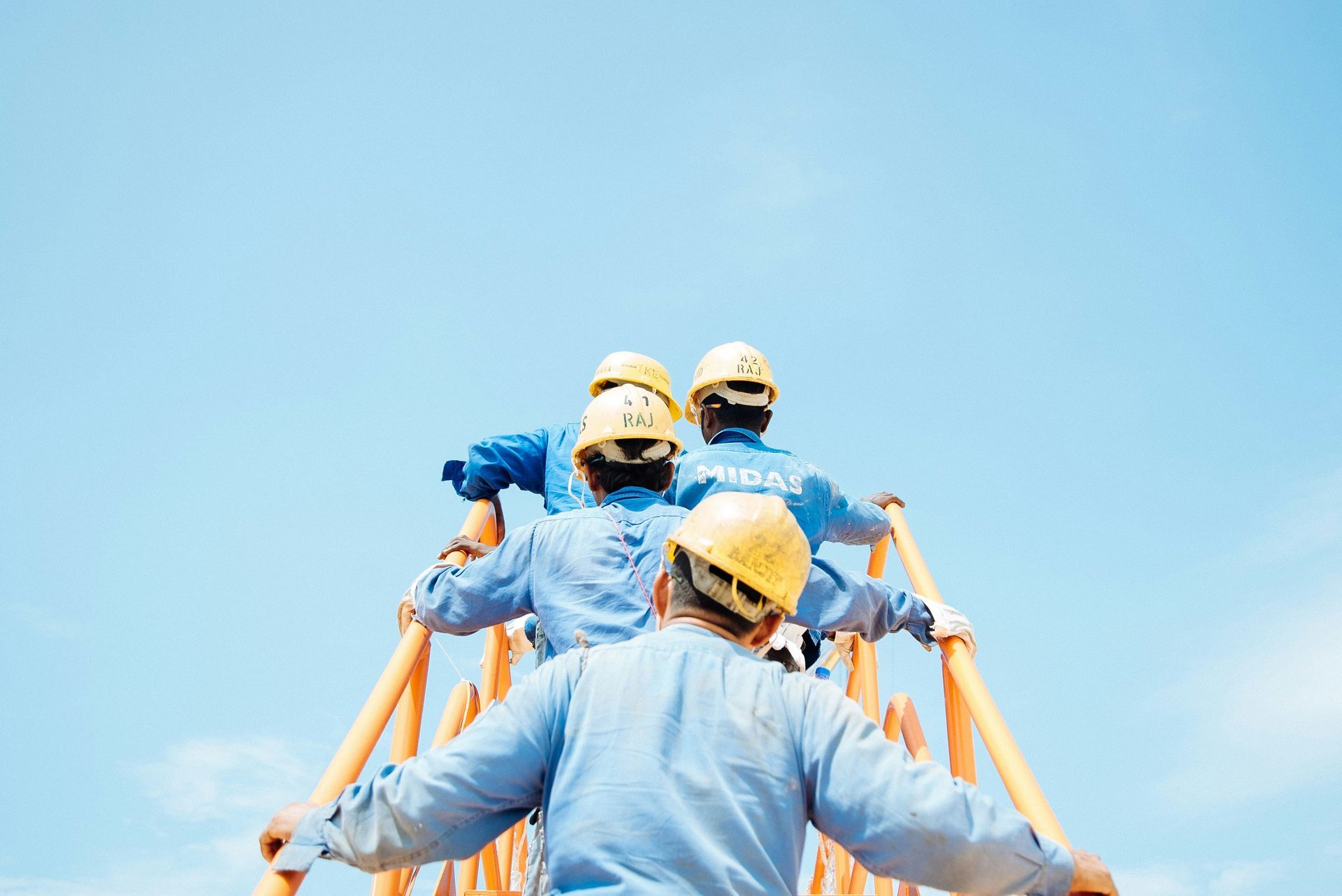 Workers in hard hats on a structure, arms outstretched, looking up.