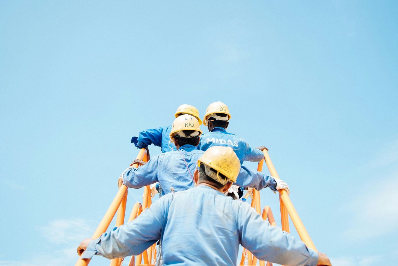 Workers in hard hats on a structure, arms outstretched, looking up.