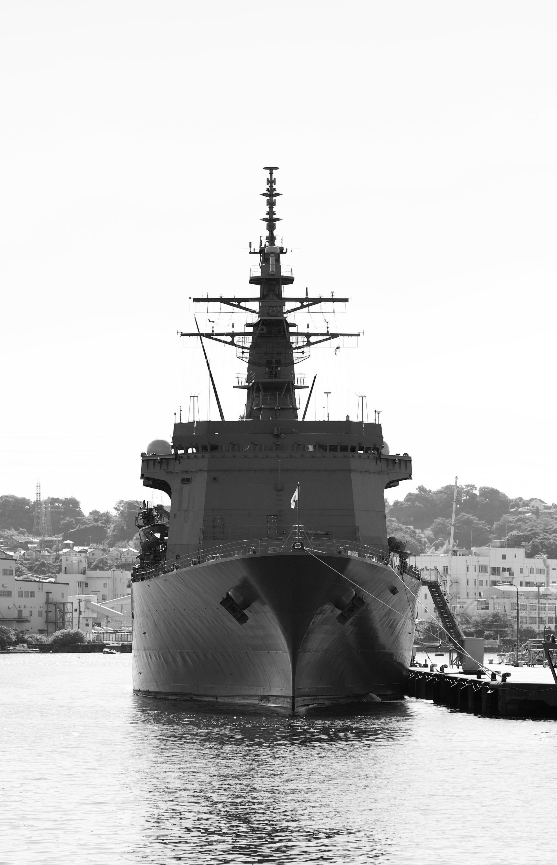 A naval ship docked at a pier, viewed head-on with antenna tower visible, houses and sky in the background.