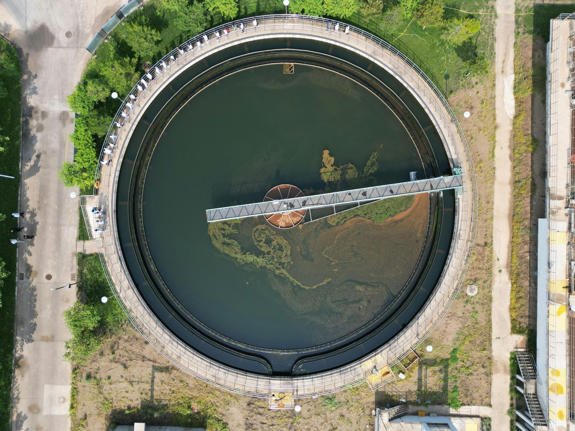 Overhead view of a large, circular water treatment tank with dark water, surrounded by concrete and vegetation.