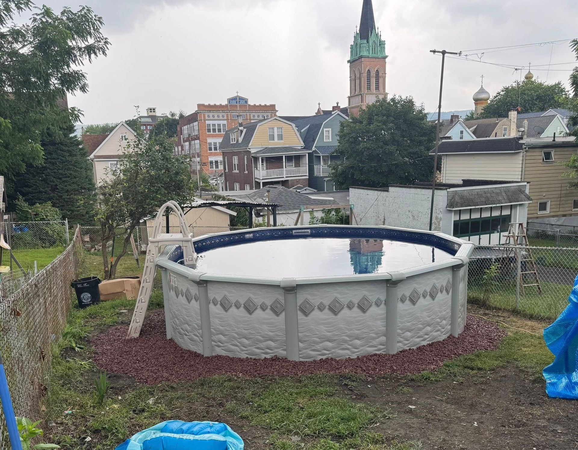 Above-ground pool in a yard, surrounded by landscaping and a view of a city with a church steeple.