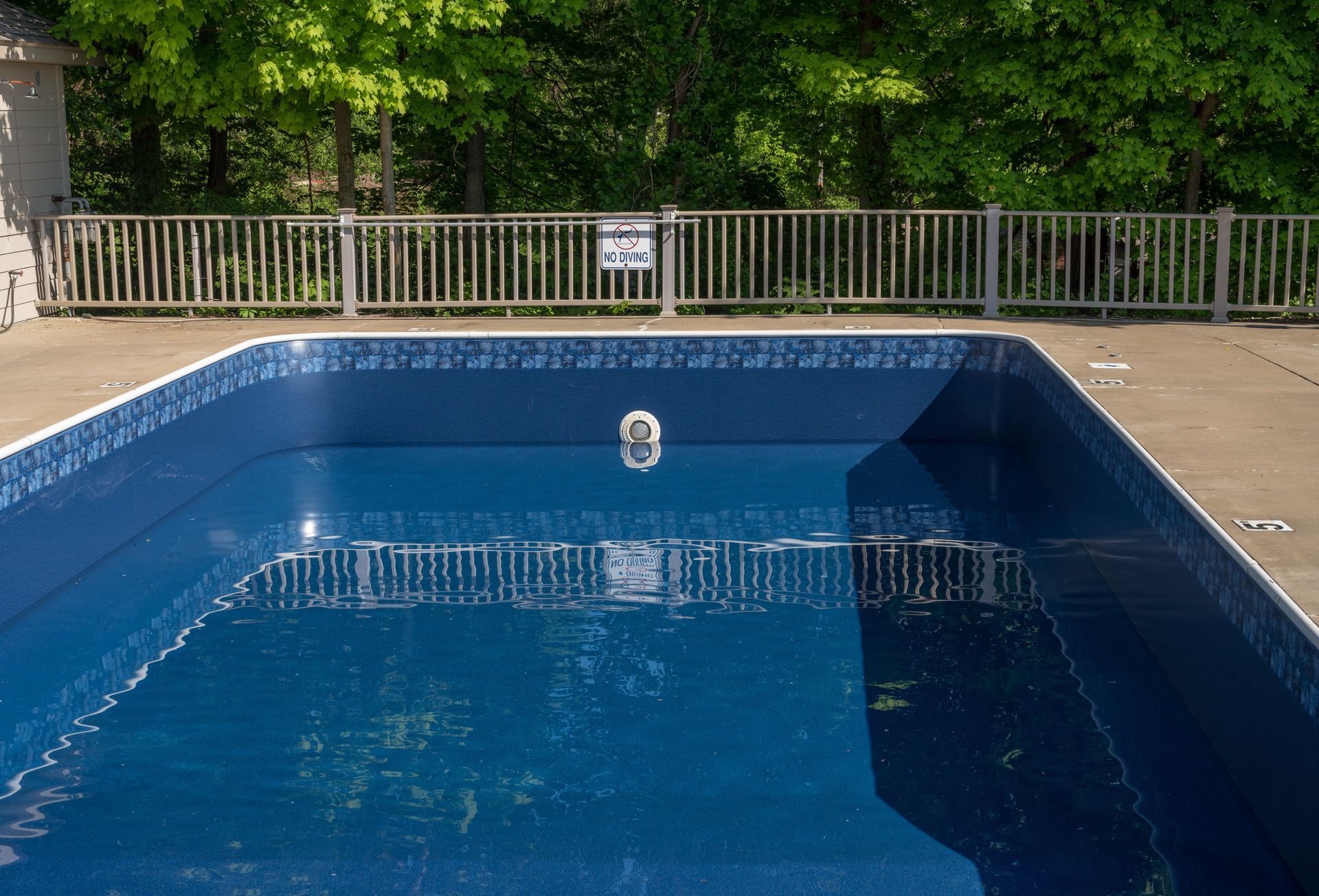 Empty rectangular pool with a blue liner, surrounded by a gray concrete deck and a metal fence. Trees are in the background.