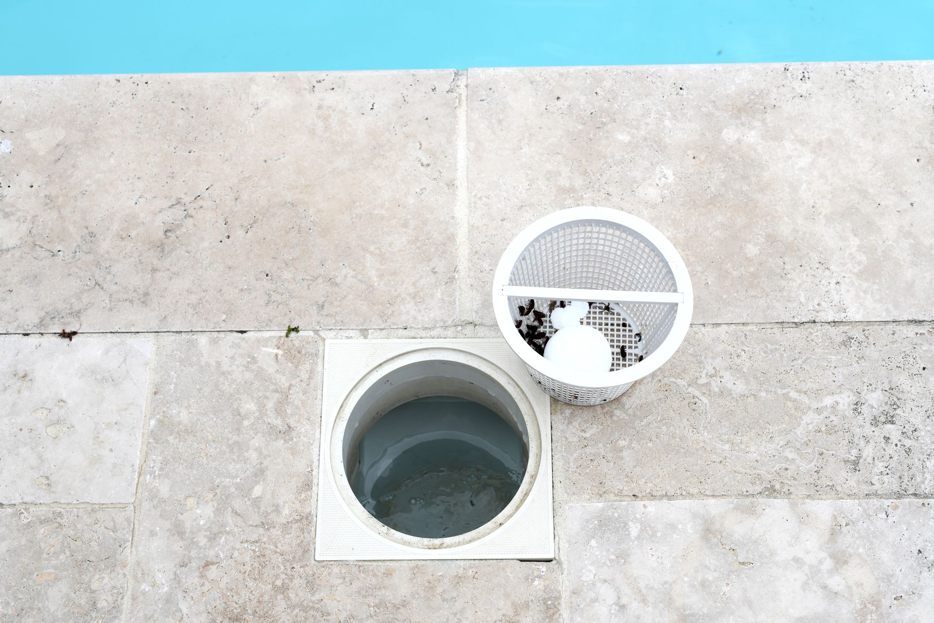 White pool skimmer basket beside its opening in a beige stone deck, with blue pool water in the background.