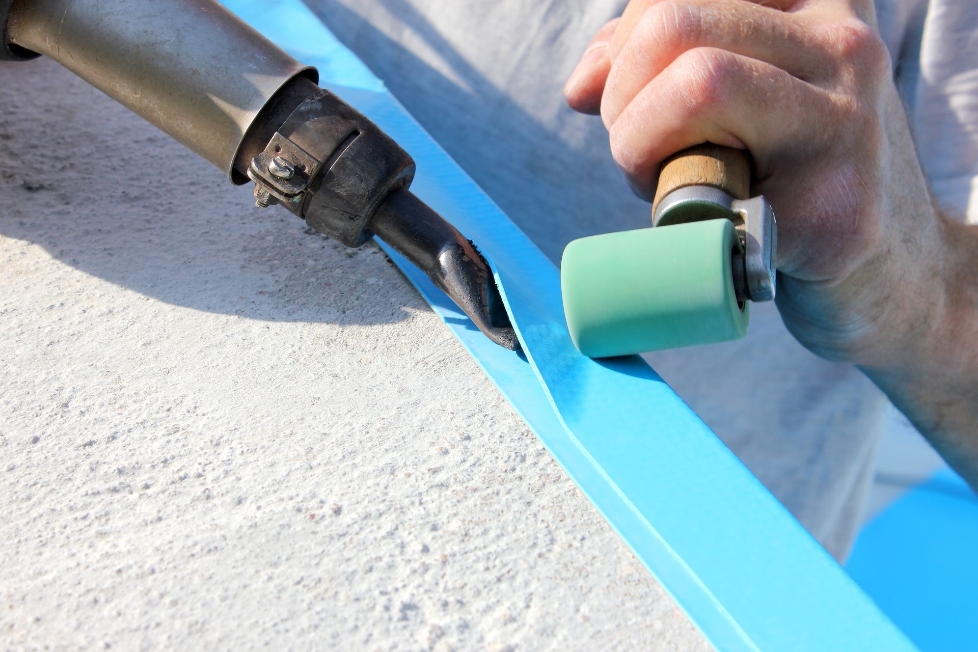 A person welds blue plastic roofing with a hot air gun and roller on a concrete surface.