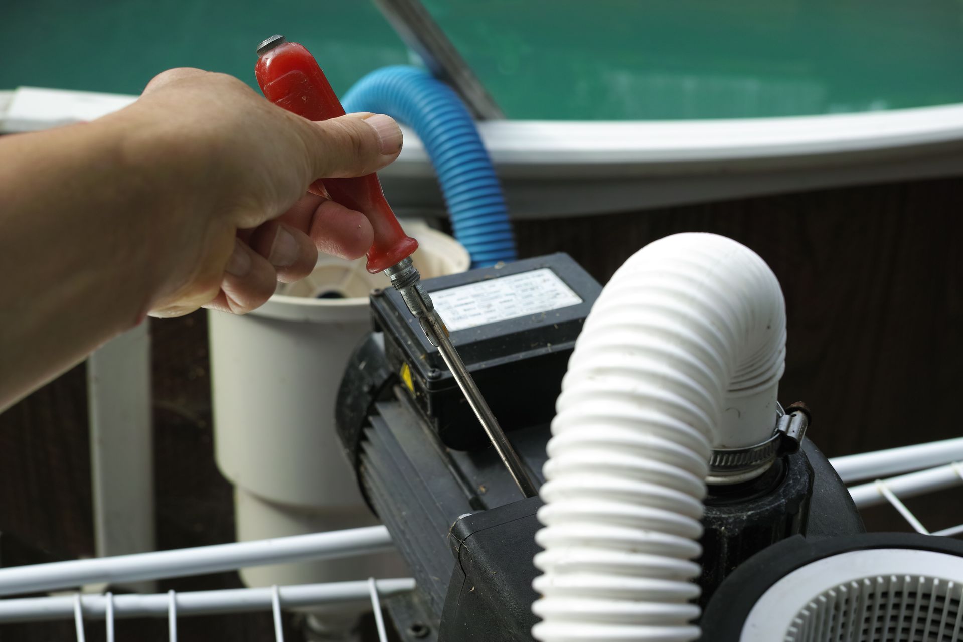 Hand using a screwdriver to repair a pool pump next to a pool.