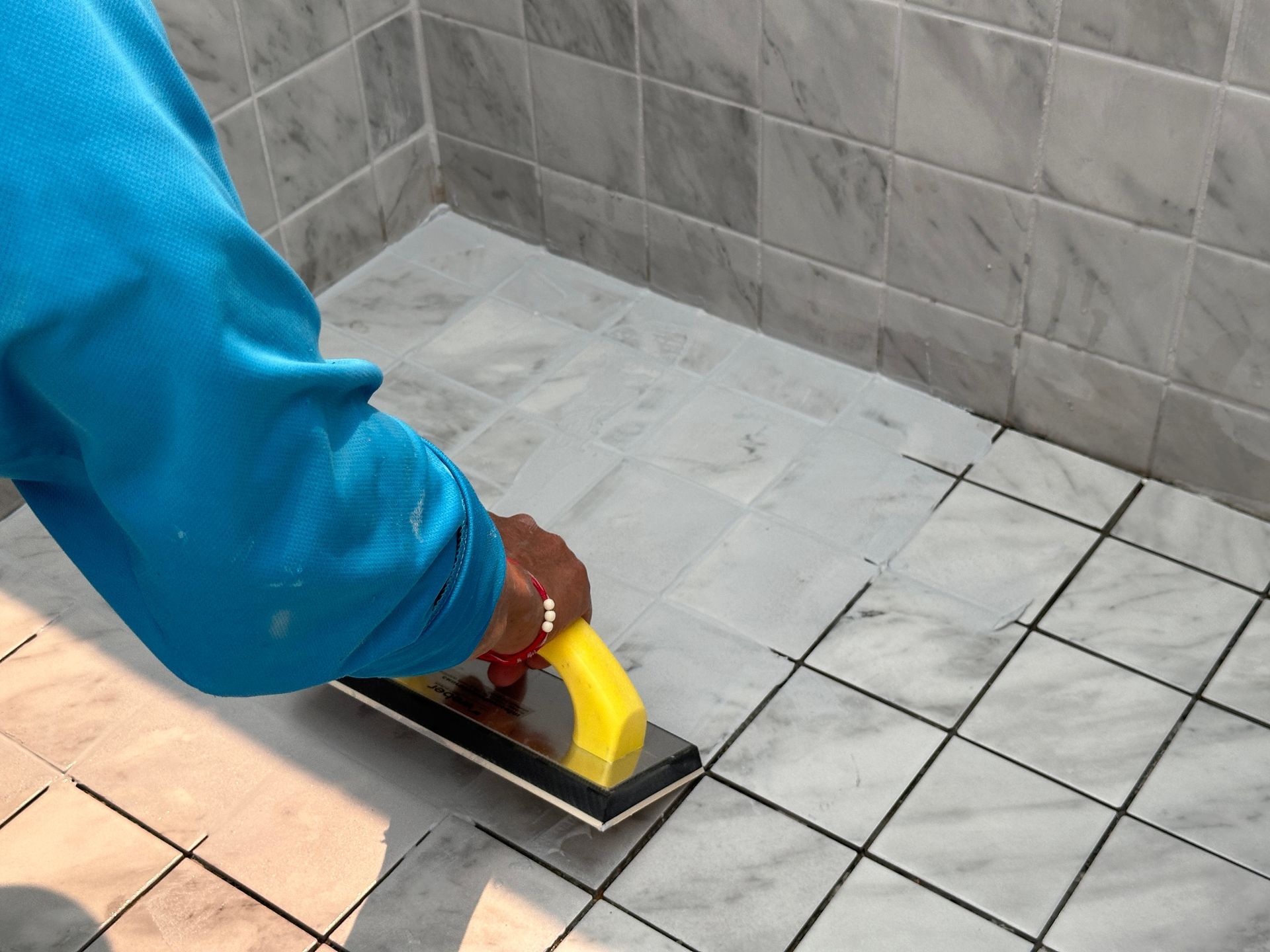 Person in a blue shirt using a grout float to apply grout to a tiled shower floor.