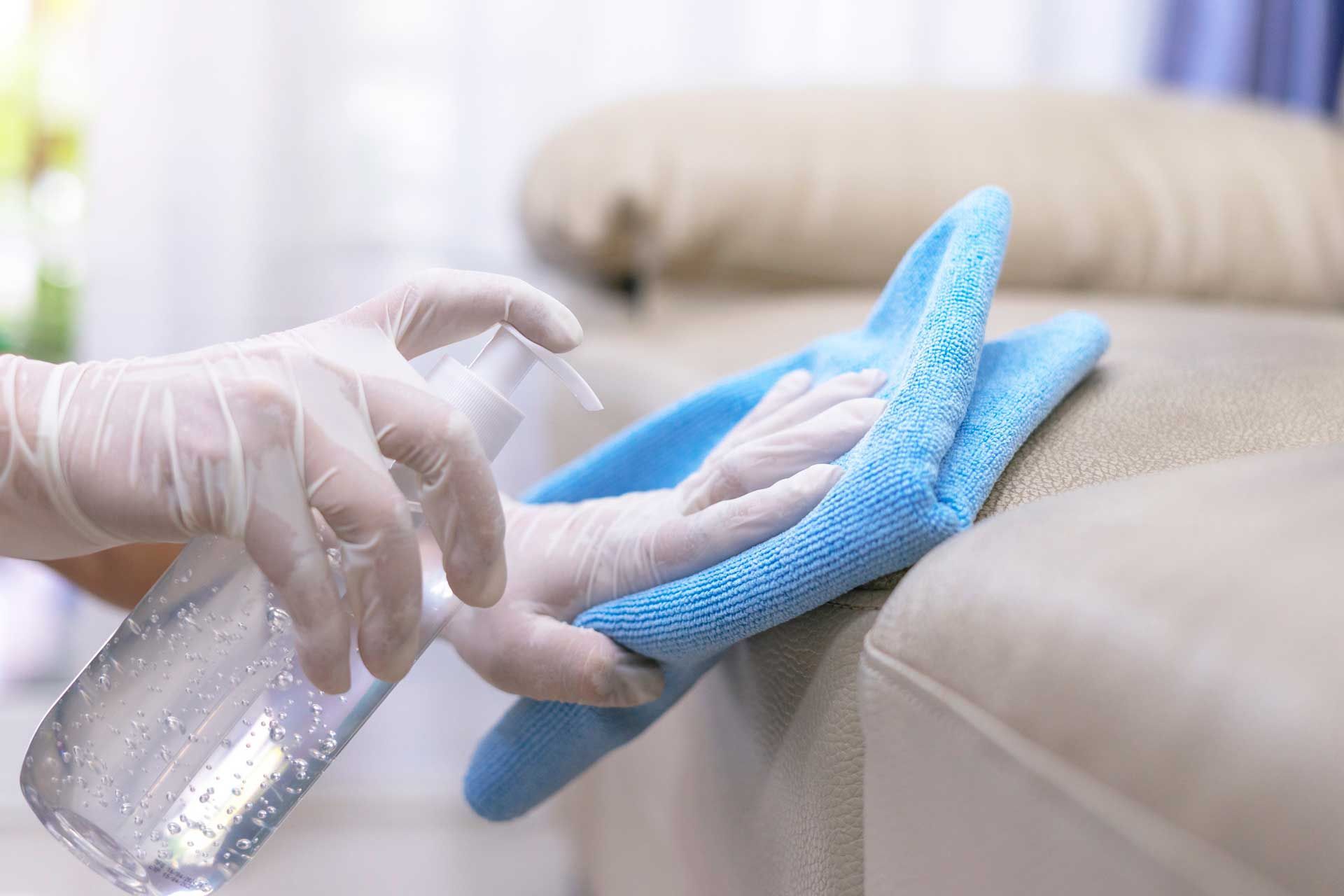 Person wearing gloves spraying and wiping down a beige sofa with a blue cloth.