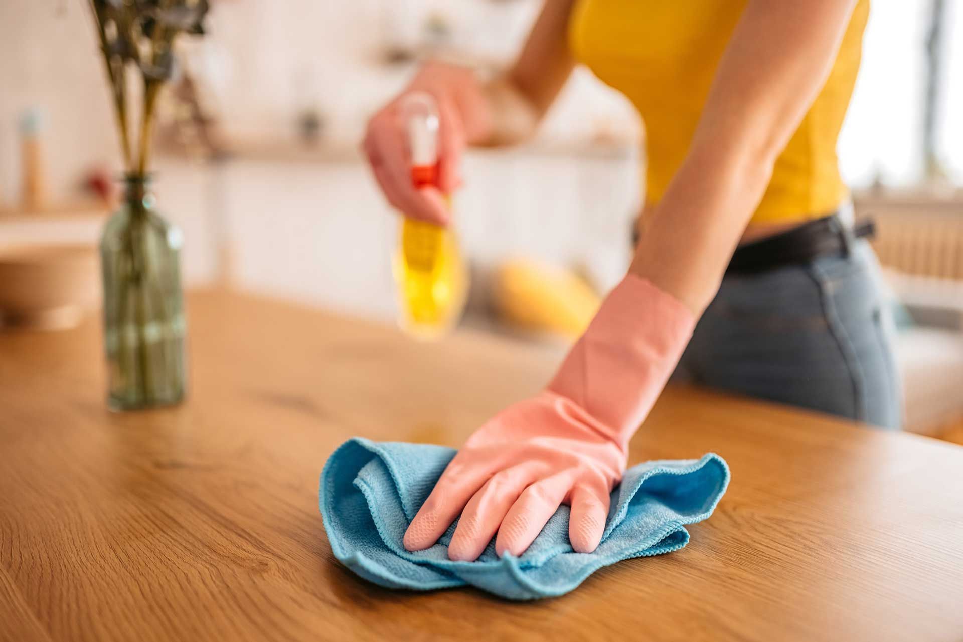 Woman wearing pink gloves cleaning wooden surface with blue cloth and spray bottle.