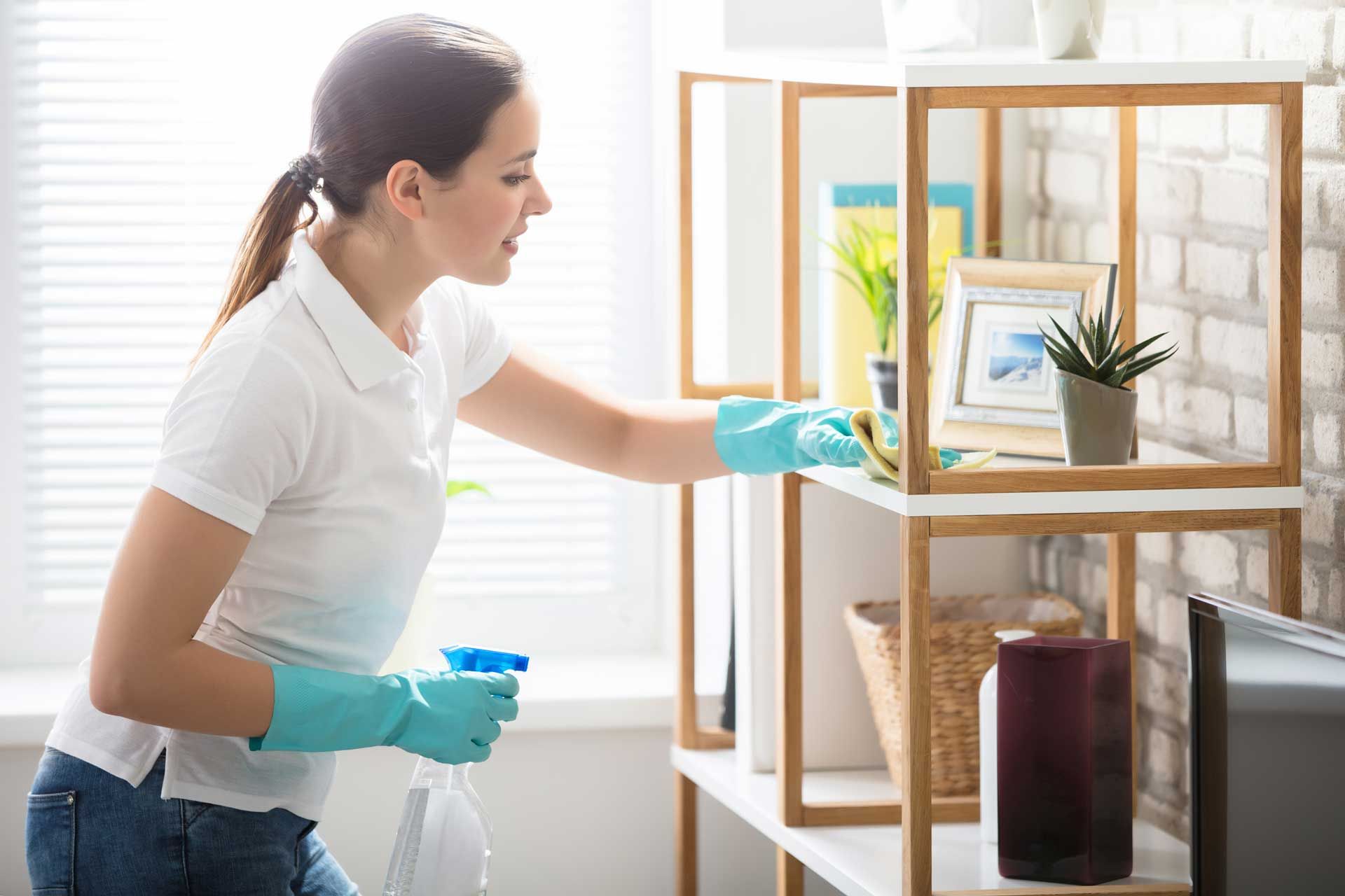 Woman wearing gloves cleaning a wooden shelf unit, holding spray bottle.