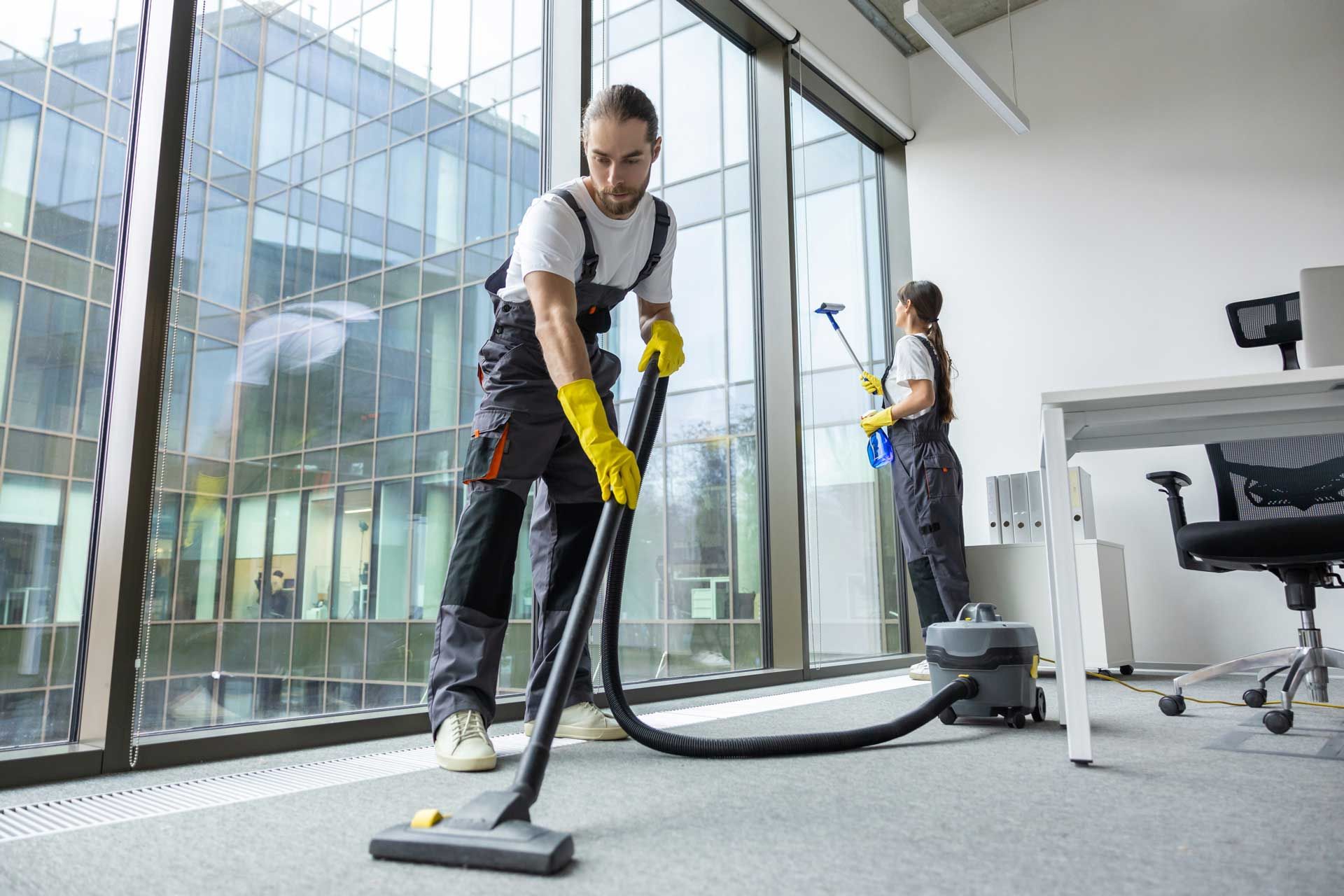 Two people cleaning an office: one vacuuming the carpet, the other dusting. Large window in the background.