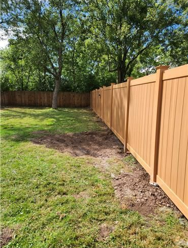 Brown vinyl fence lines a grassy backyard. Green trees are in the background.