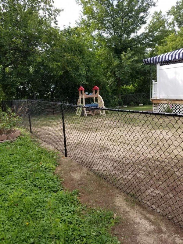 Black chain-link fence encloses a yard with a playset. Green grass, trees, and a house are in the background.