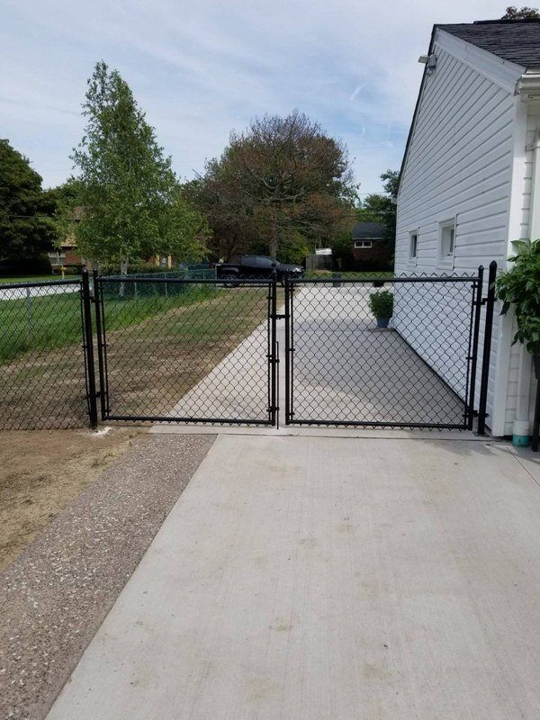 Black chain-link fence gates at the end of a concrete driveway, next to a white house.