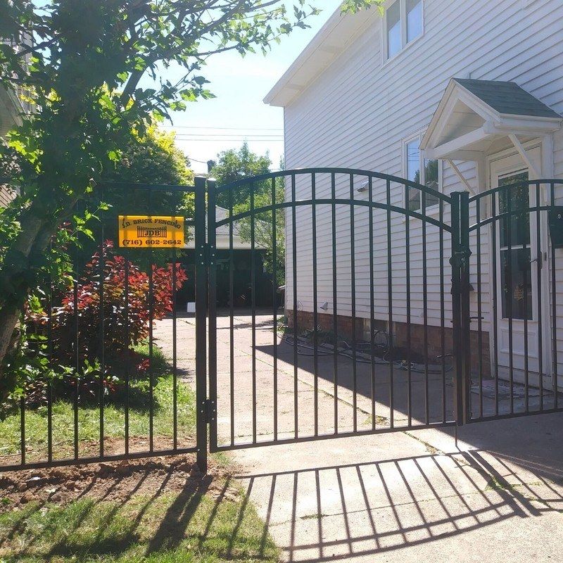 Black metal gate in front of a white house, with a sign. Sunlight casts shadows on the concrete driveway.