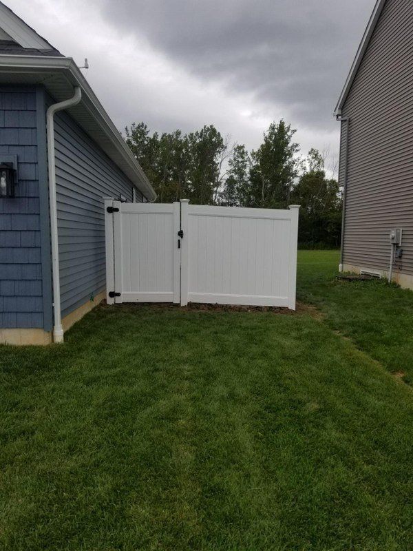White vinyl fence with gate between two houses on a grassy lawn under a cloudy sky.