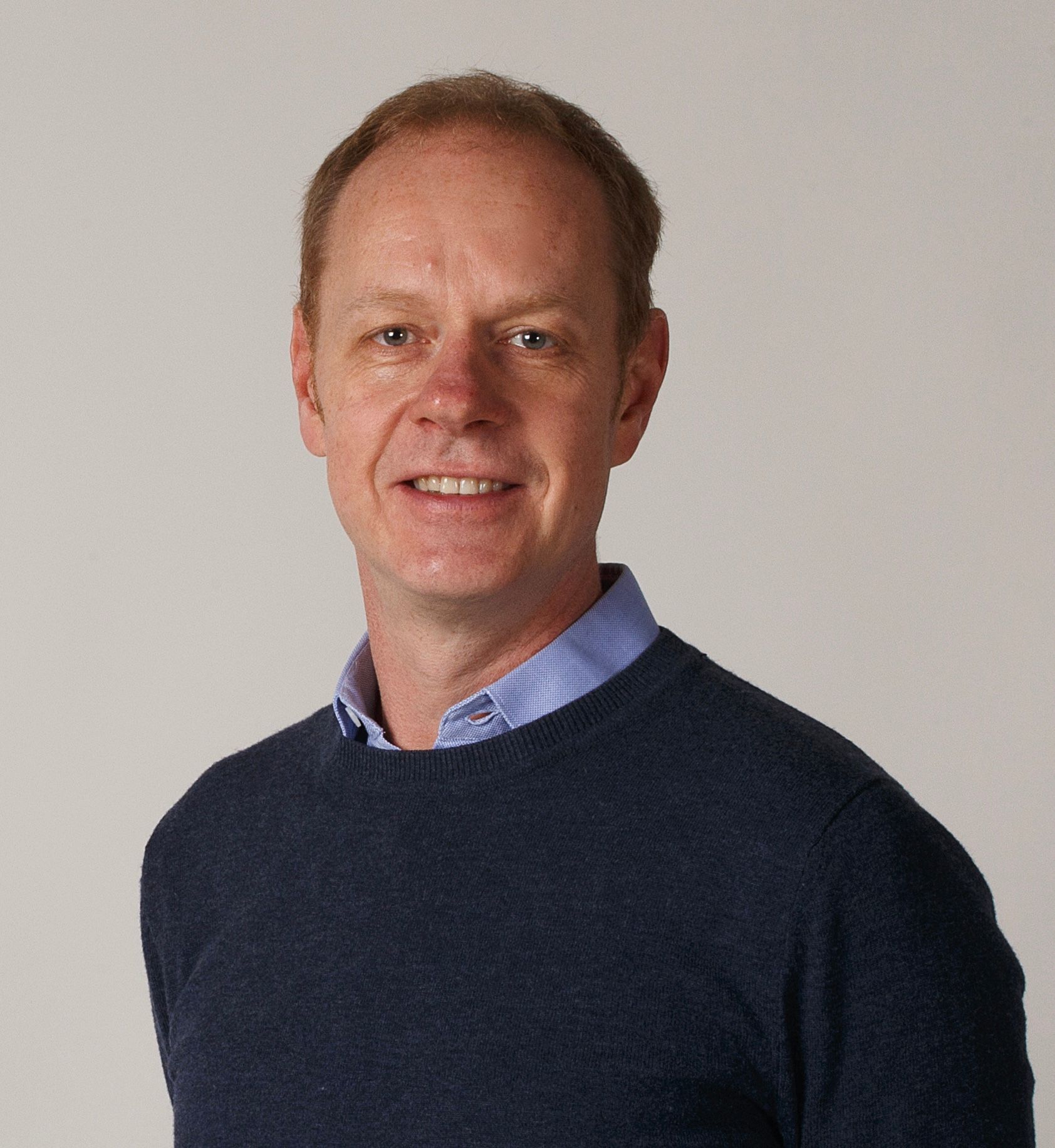 Hymyilevä silmälasipäinen mies, jolla on musta poolopaita, harmaata seinää vasten.