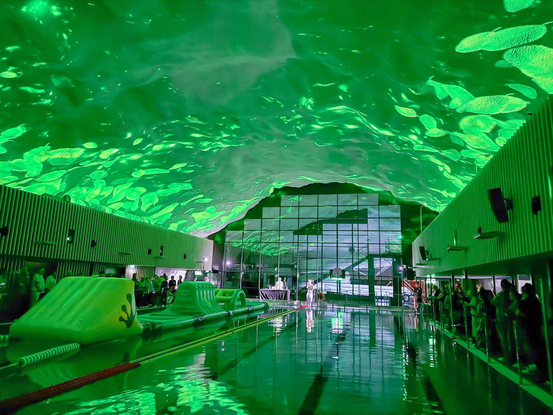 Sisäuima-allas, jossa on vihreä valaistus ja puhallettavia esteitä. Vesi heijastaa valoja. Ihmiset seisovat reunoilla.