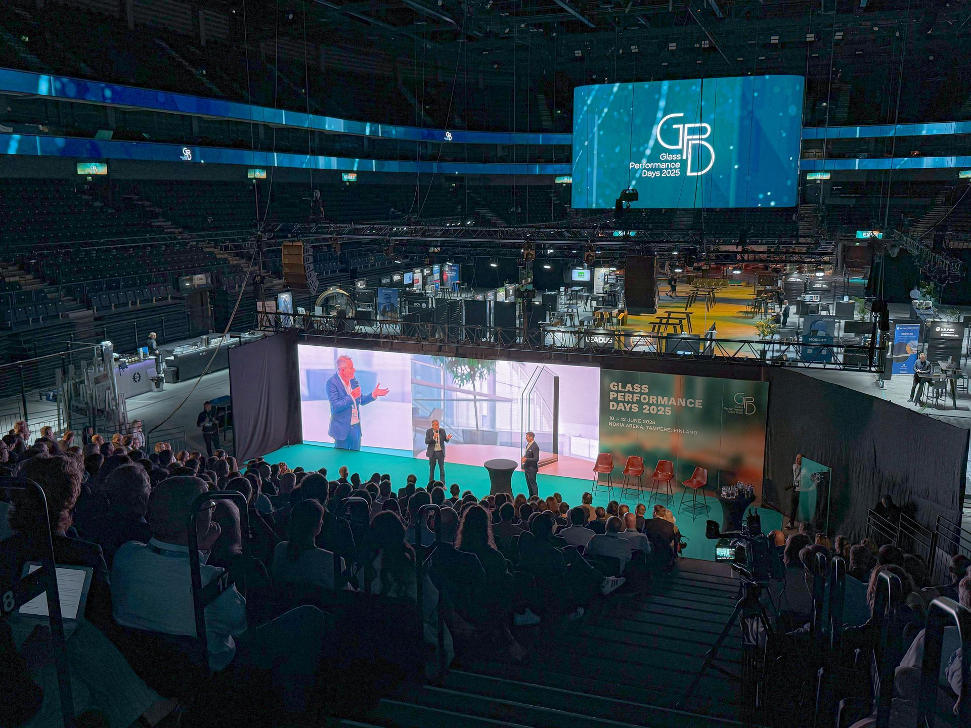 Yleisö konferenssissa areenalla; puhujat lavalla näyttöjen edessä; lavabanneri: