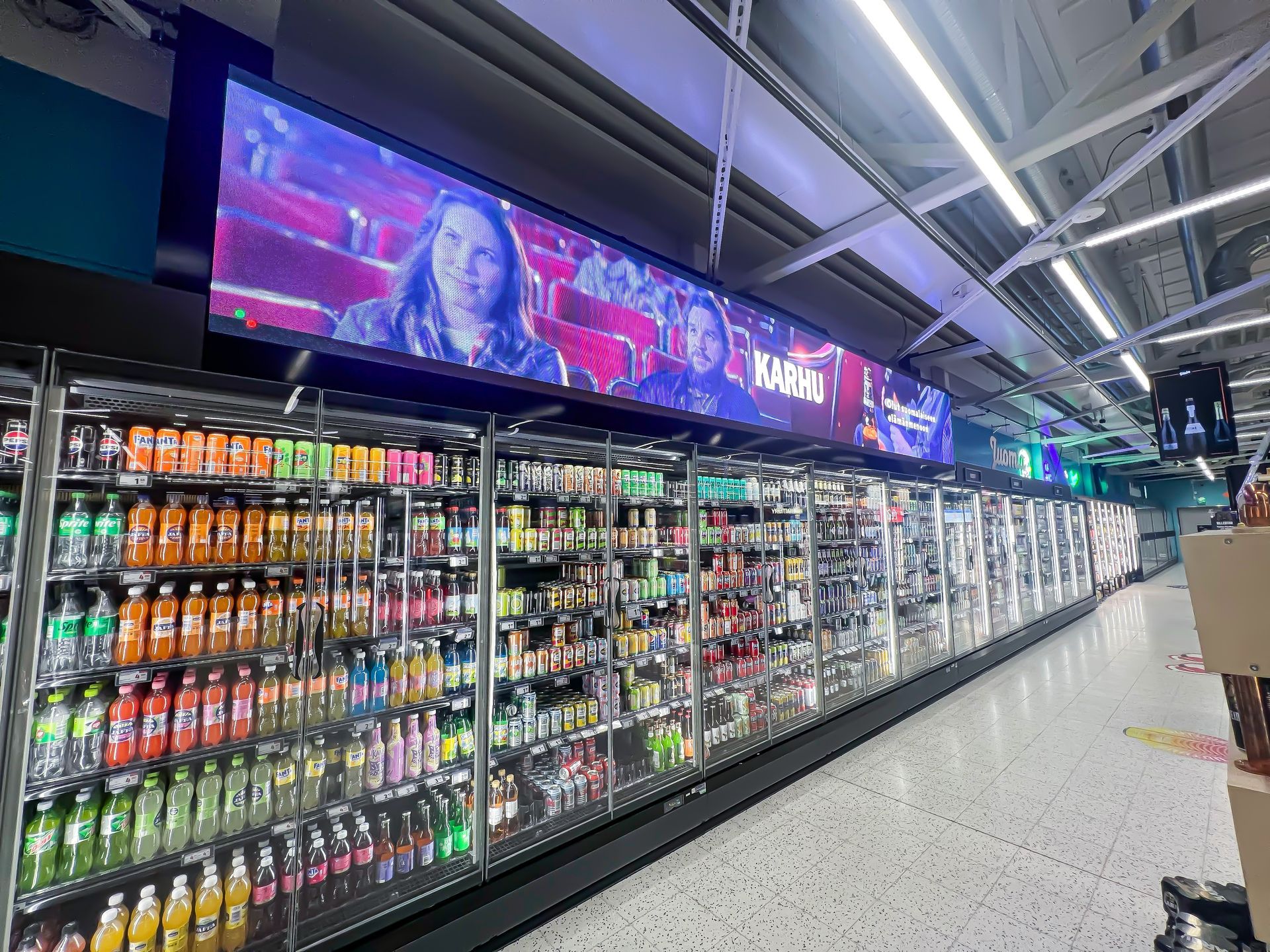 Kirkkaasti valaistu ruokakaupan juomahylly, jonka digitaalinäyttö näyttää mainoksen kylmähyllyjen yläpuolella.