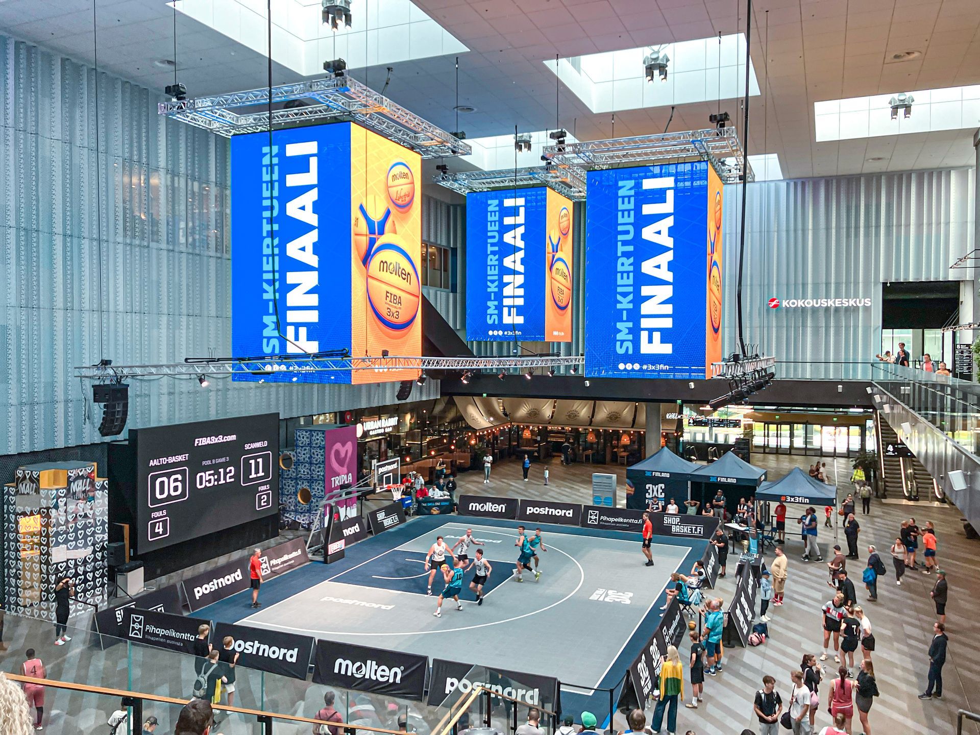 Koripallo-ottelu suuressa atriumissa; pelaajat kentällä, tulostaulu näkyvissä. Banderolleissa lukee 