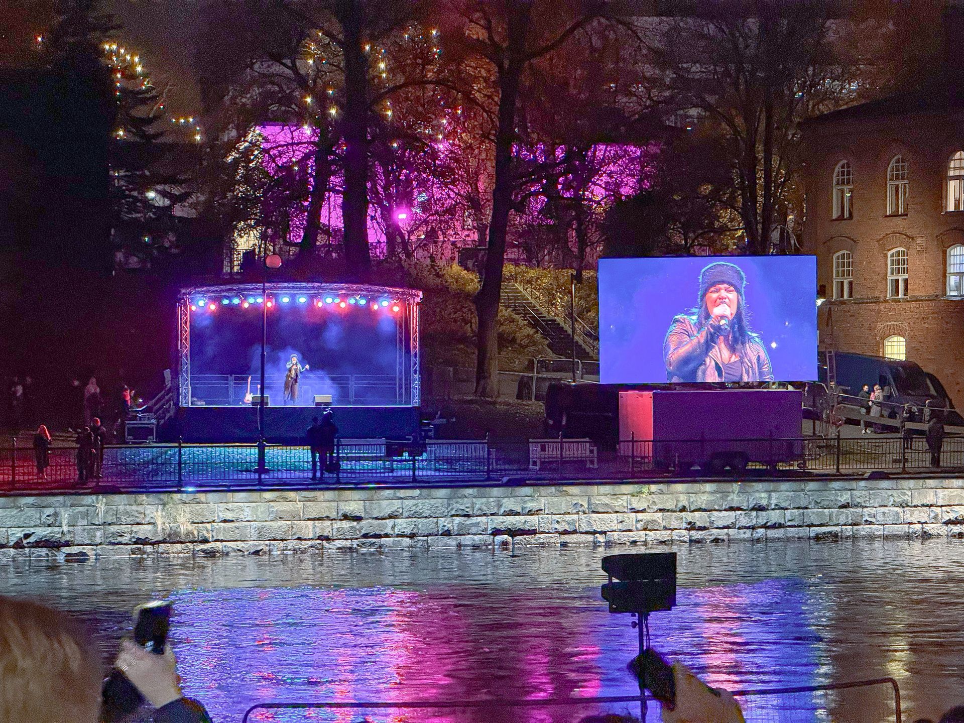 Vedessä oleva lava, jossa on esiintyjä ja suuri, värikkäillä valoilla valaistu näyttö, joka näyttää esityksen.