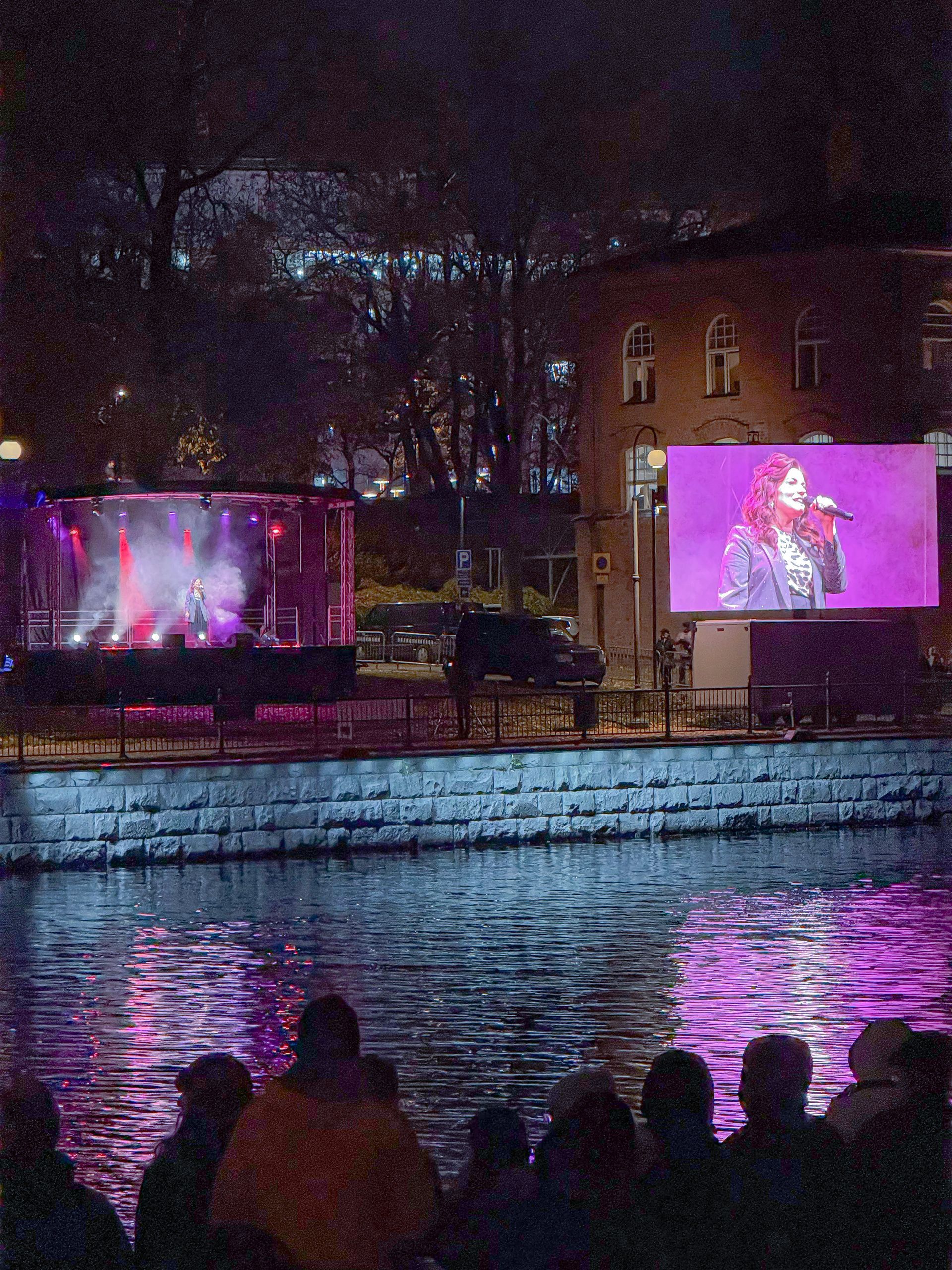 Live-konsertti yöllä. Esiintyjä laulaa lavalla, ja hänen äänensä näkyy veden yllä olevilla suurilla näytöillä.