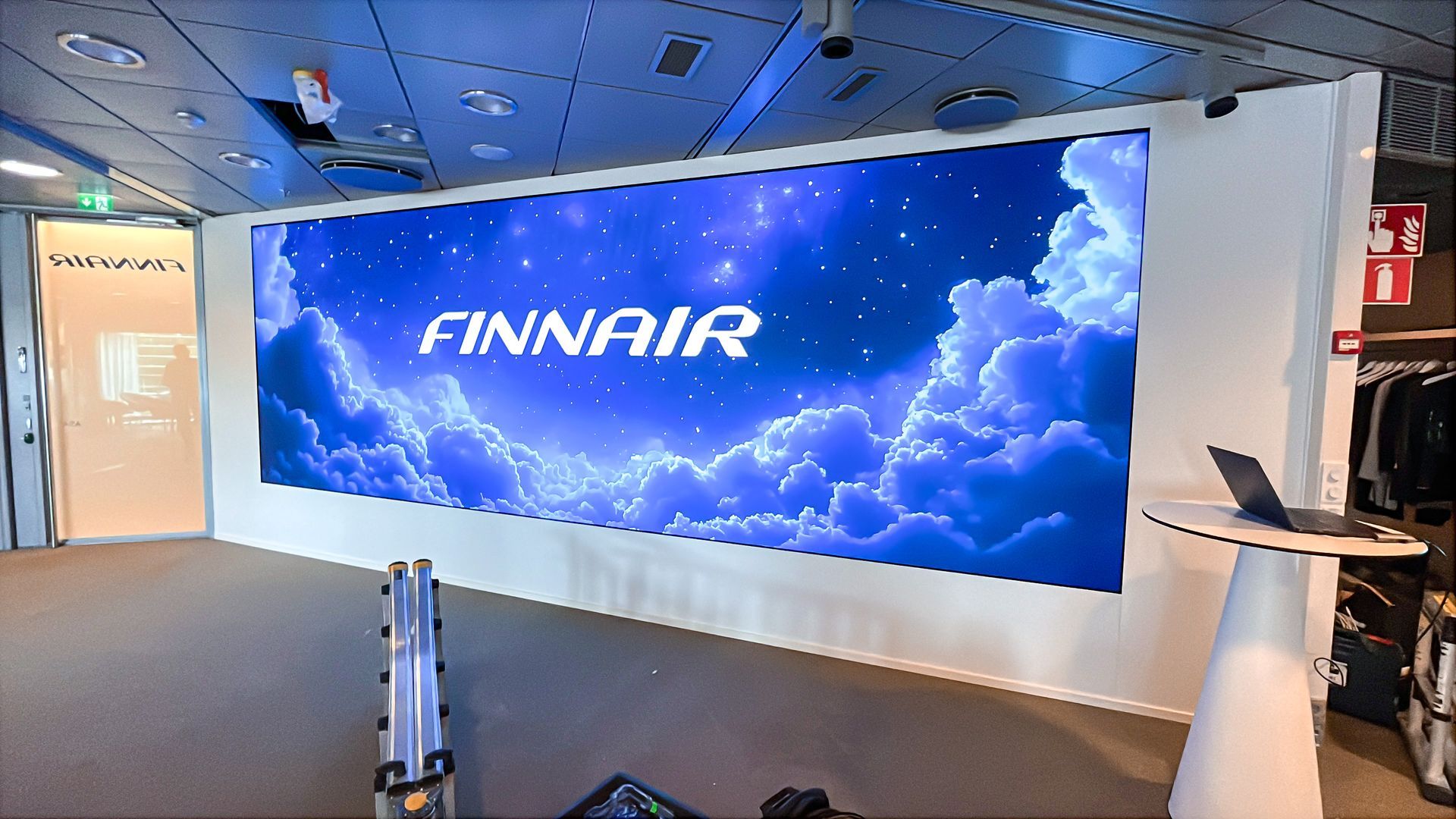 Suuri näyttö, jolla näkyy Finnairin logo pilvistä taustaa vasten. Sisätilat pöydällä ja kannettavalla tietokoneella.