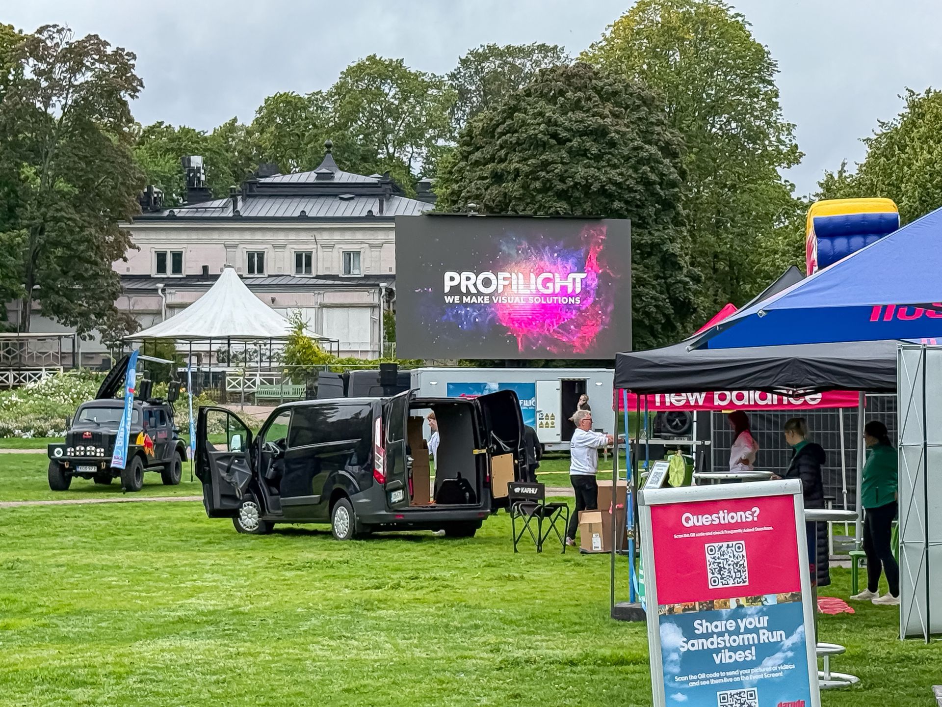 Tapahtuma-asetelma puistossa, jossa on näyttö, pakettiautoja, telttoja ja ihmisiä.