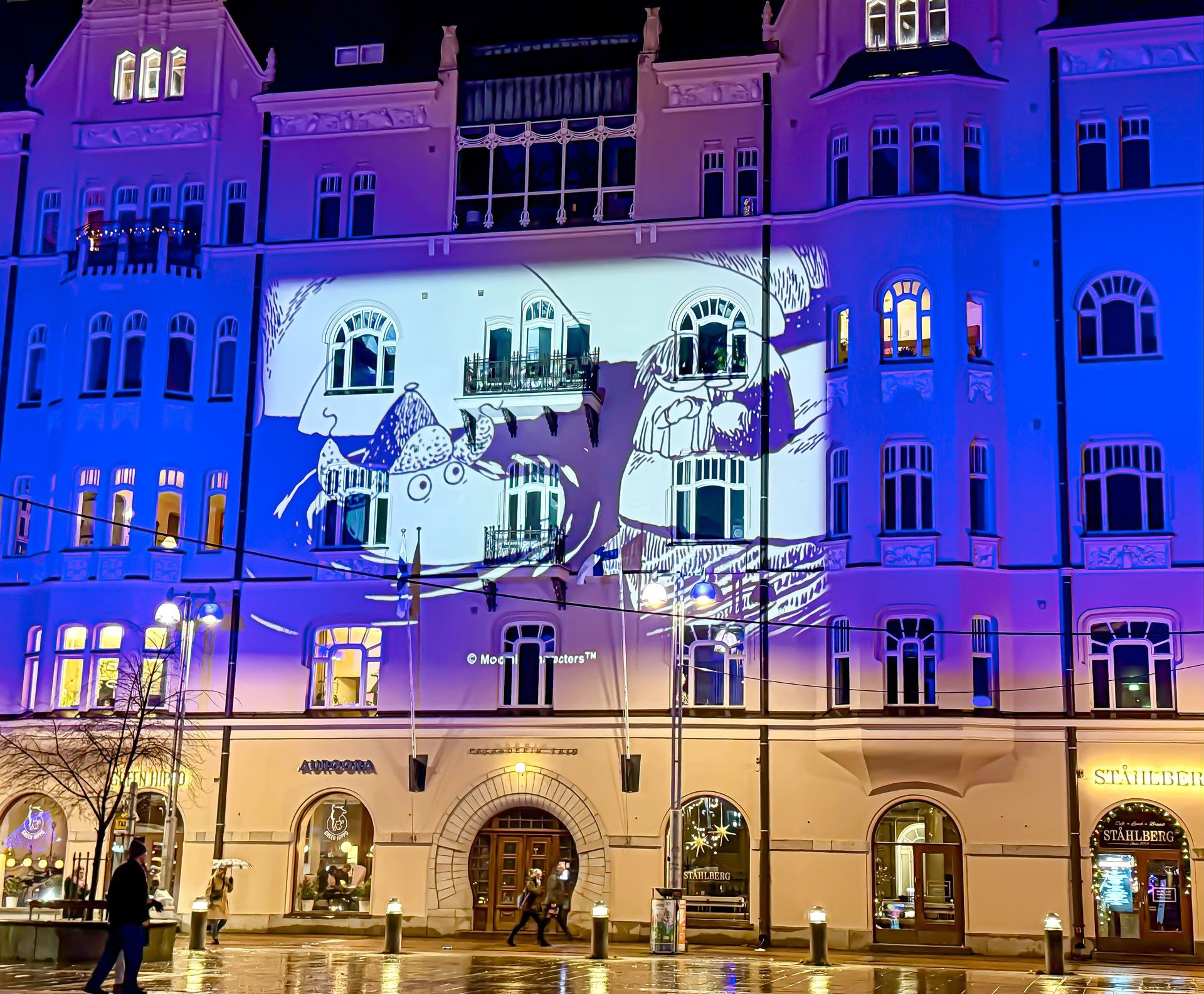 Rakennuksen julkisivu yöllä, jossa heijastettu valkoinen ja sininen valoshow korostaa arkkitehtonisia yksityiskohtia.