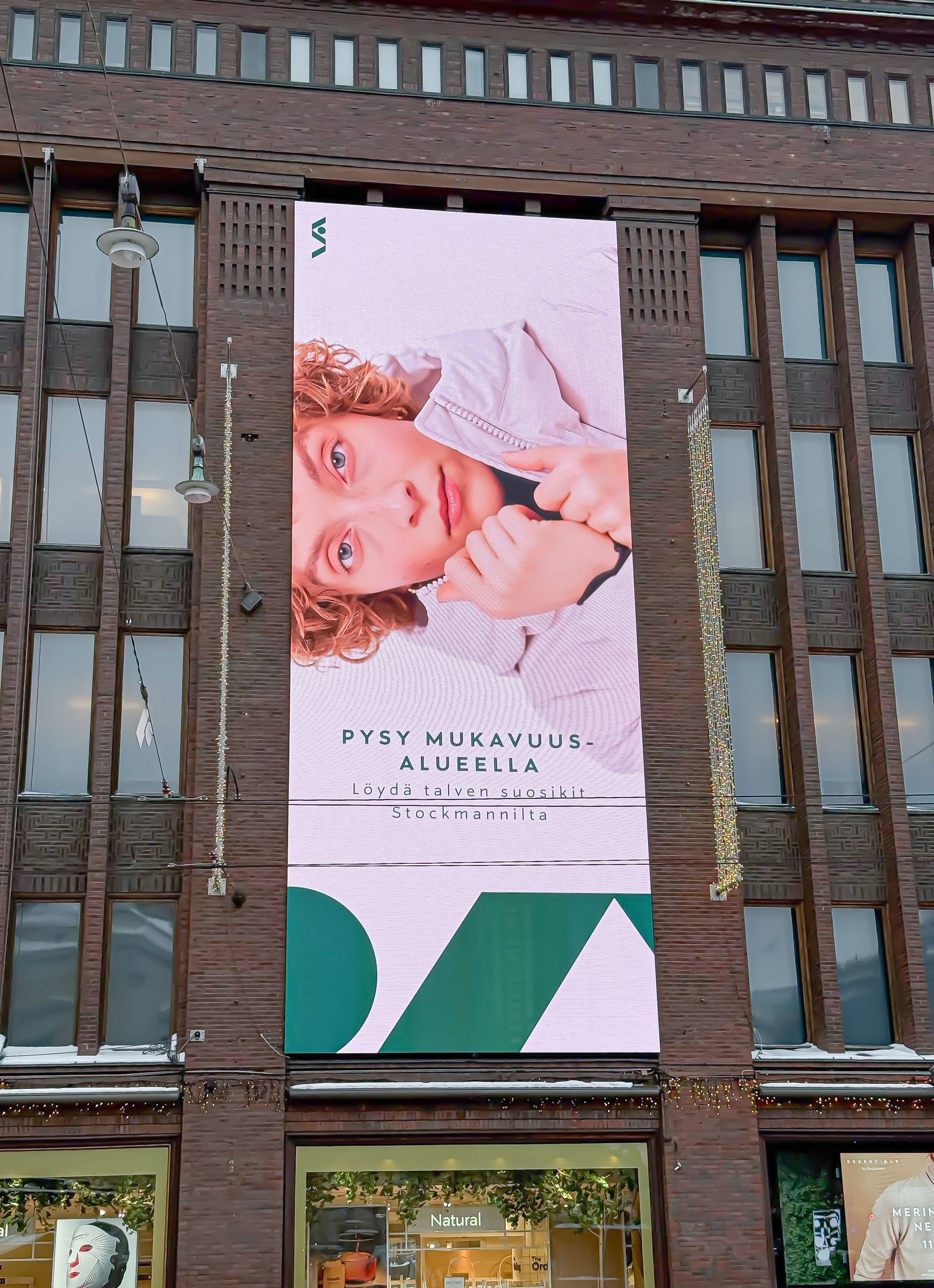 Mainos rakennuksen julkisivussa: Henkilö vaaleanpunaisessa ylhäällä, vaaleanpunainen digitaalinen tausta, vihreät sisustuselementit.
