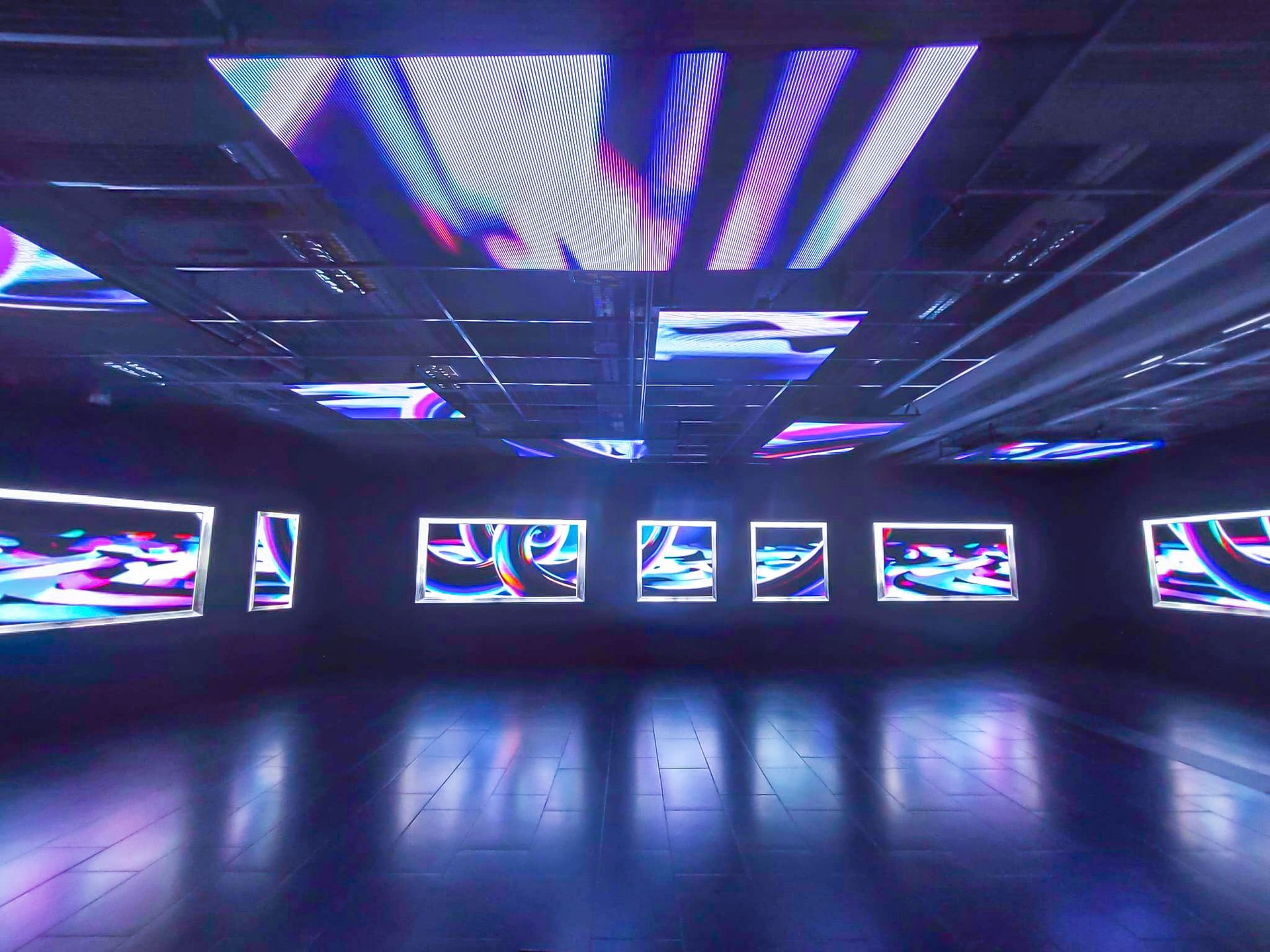 Pimeä huone, jossa on digitaalisia taideteoksia; sininen ja violetti valaistus heijastuu lattiasta.