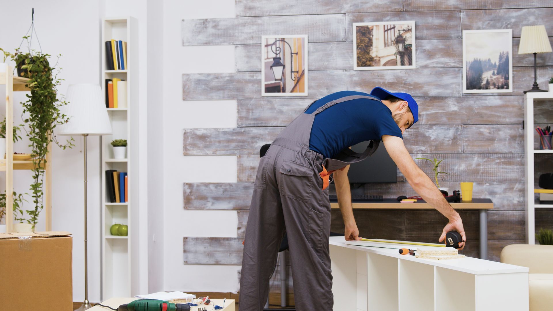 A worker in gray coveralls and a blue cap measures a white furniture unit in a living room with wooden wall paneling.