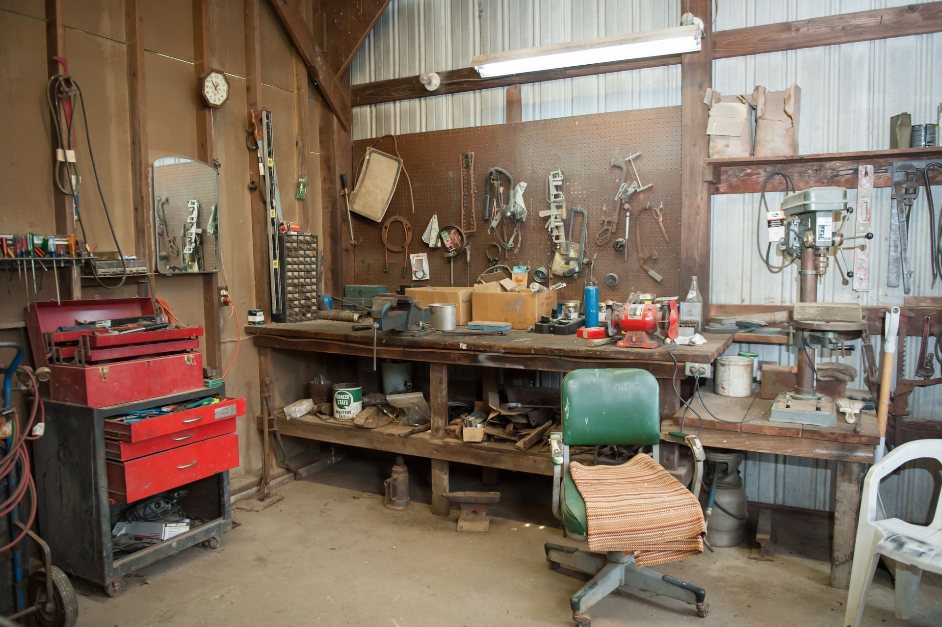 A cluttered wood-paneled workshop with a workbench, a red toolbox, tools hanging on the wall, and a green office chair.