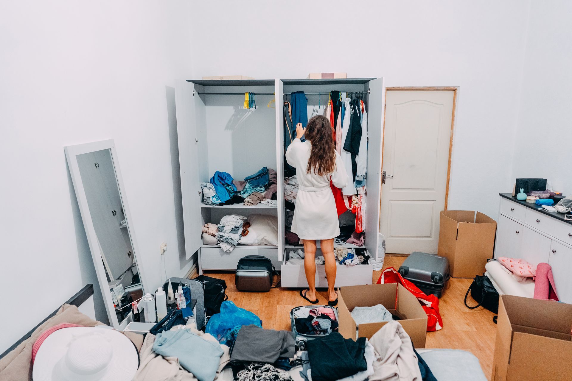 A person wearing a white bathrobe stands facing an open closet in a room filled with cardboard boxes and folded laundry.