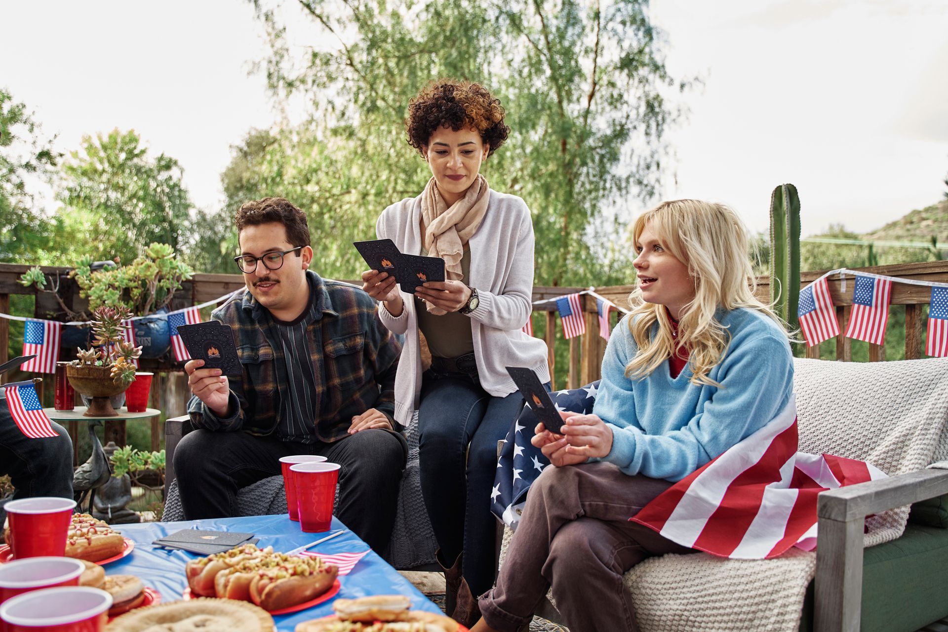Three people playing cards around a table with snacks and American flag decorations at an outdoor gathering.