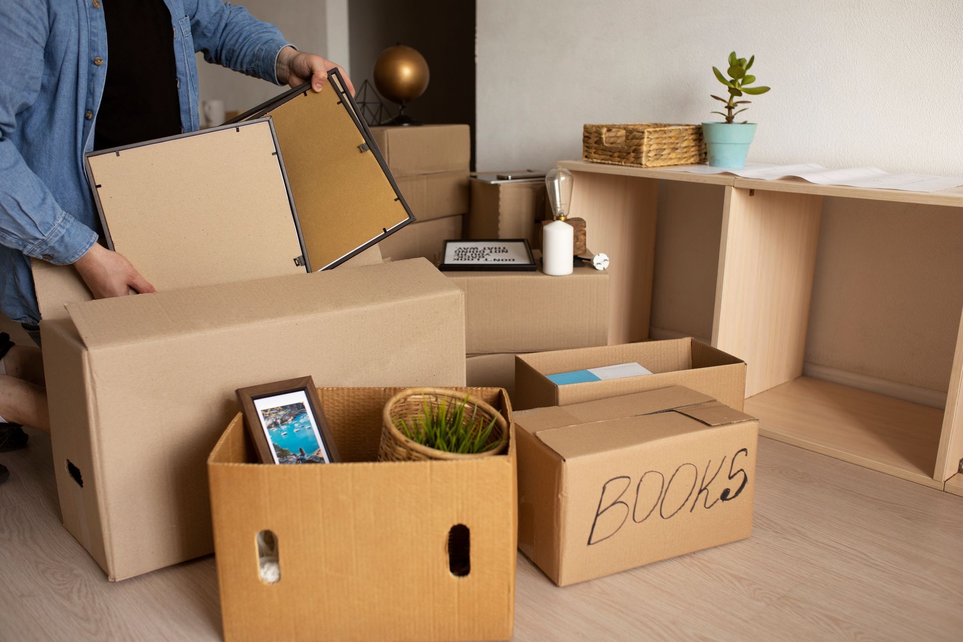 Person packing moving boxes in a bright room, placing a framed picture. Several packed boxes are visible.