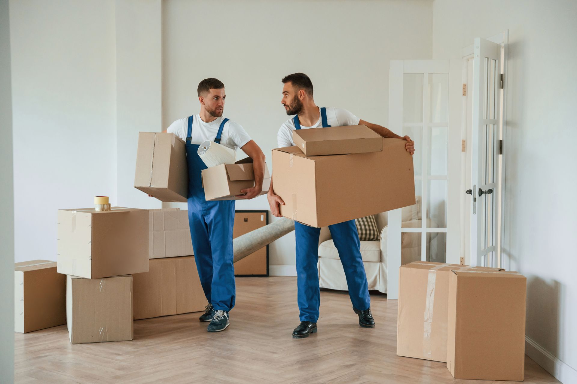 Two movers in blue coveralls carrying cardboard boxes in a bright room.