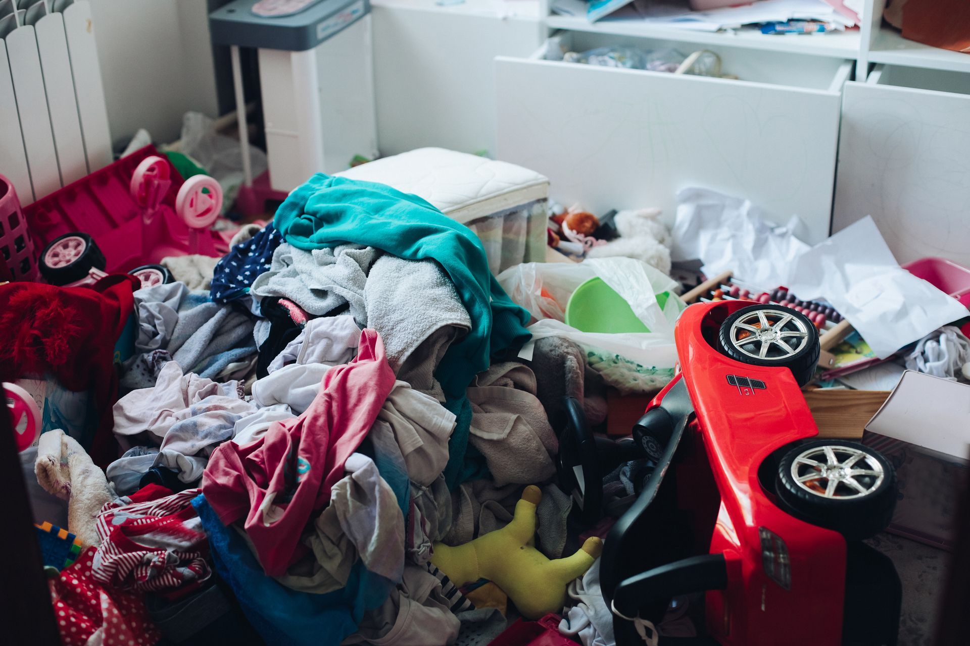 Pile of clothes and toys in a cluttered room with a red car.